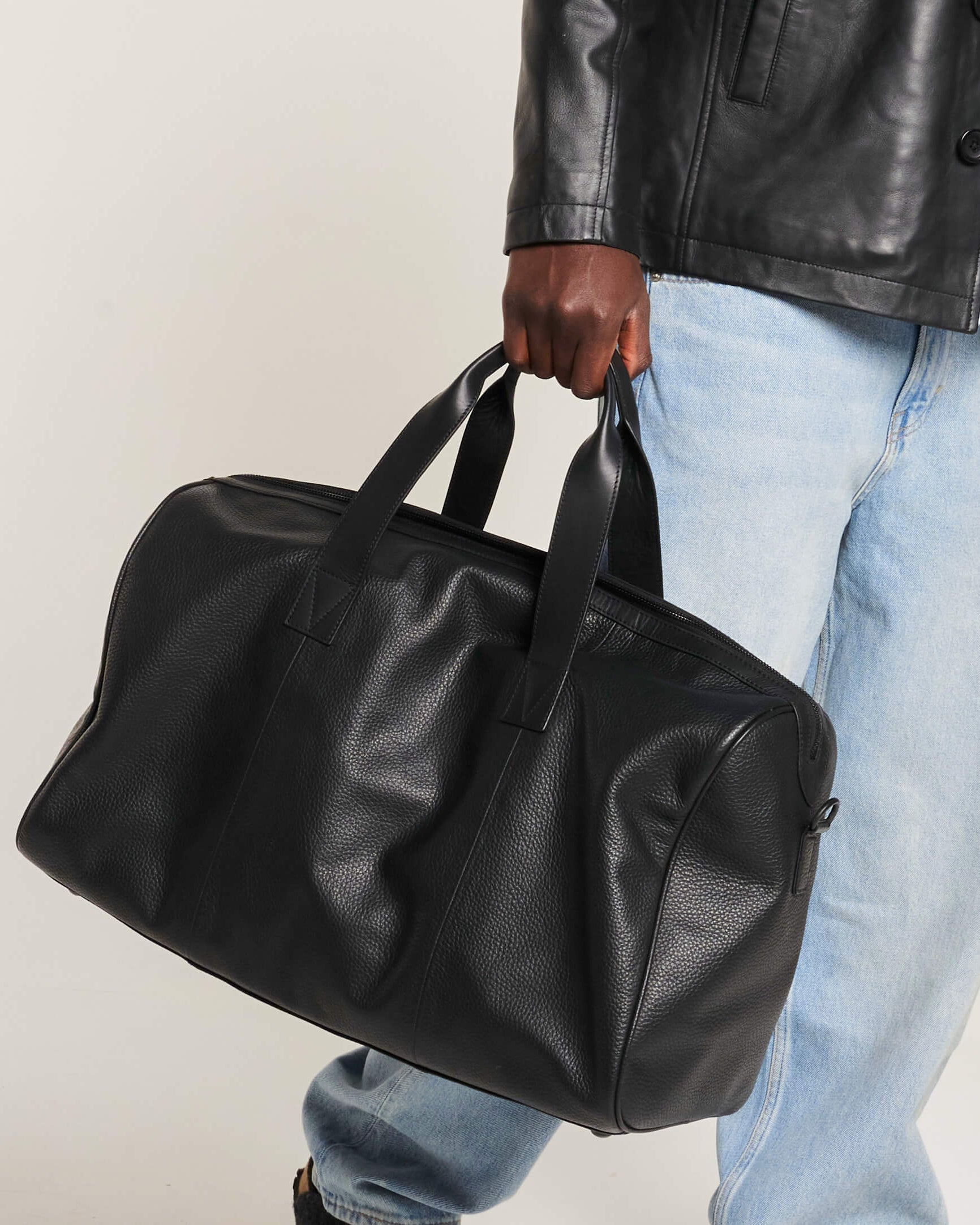 Person holding a black leather duffel bag against a neutral background