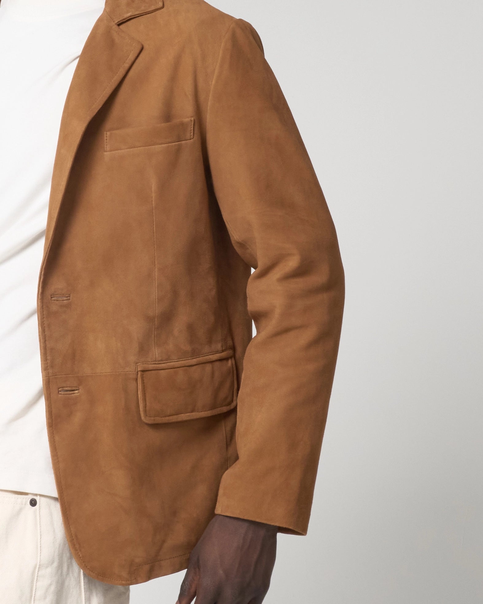 A close-up of a person wearing a tan goat suede blazer over a white shirt and light trousers. The blazer has an upper and lower pocket.