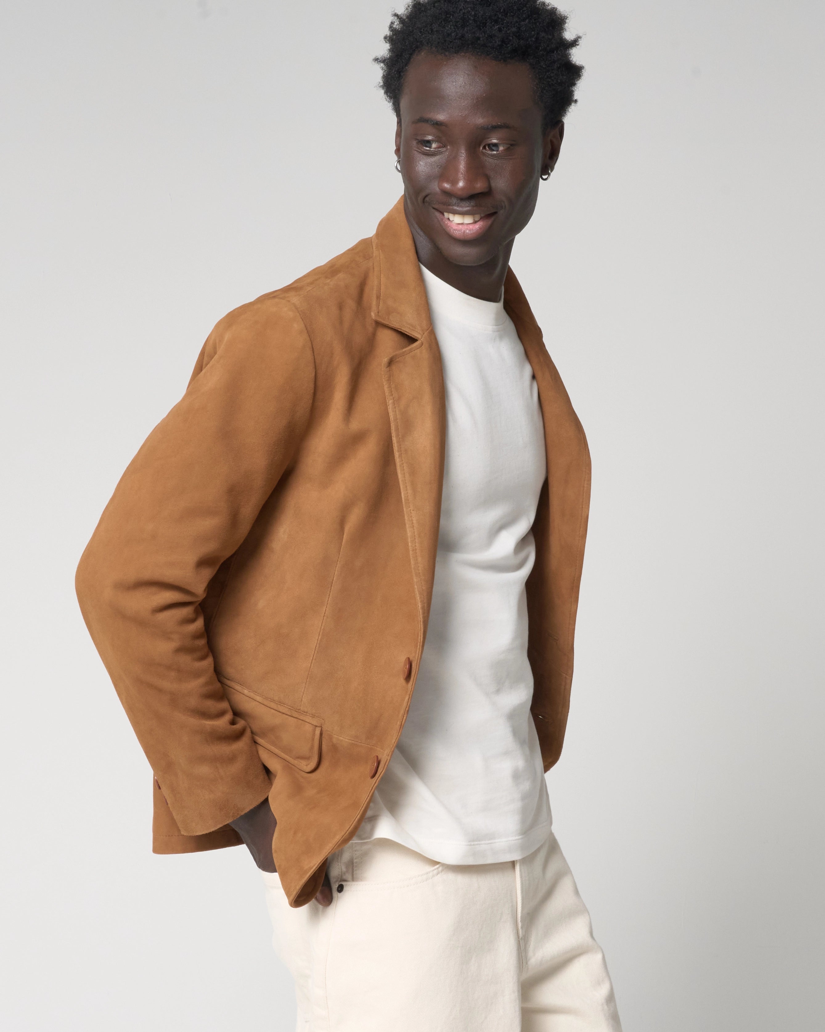 A man wearing a tan suede blazer over a white t-shirt and off-white trousers. He puts his hands in the pockets of his jeans to show how the suede jacket moves. It is smooth in appearance with no wrinkles.
