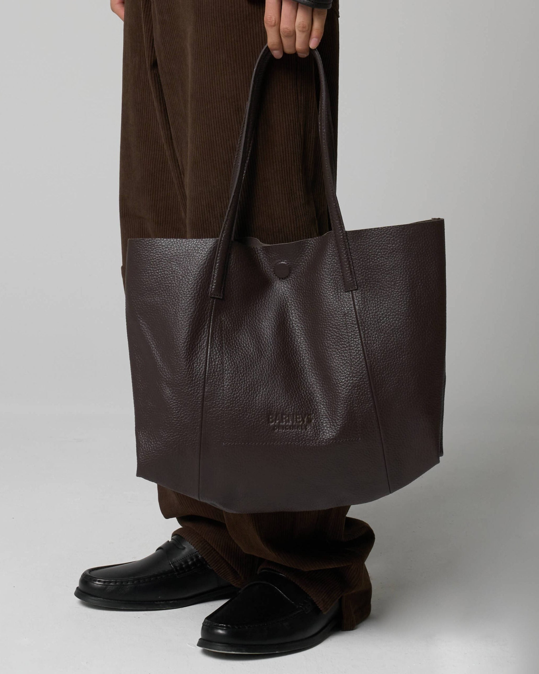 Person holding a brown leather tote bag against a plain background. The leather has a wrinkled texture and is embossed with the words 'Barneys Originals'.