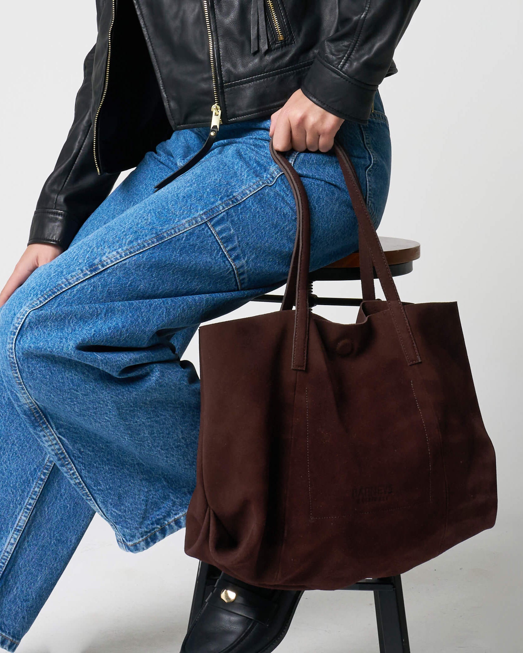 Person wearing a black leather jacket, blue jeans, and black shoes, holding a brown suede tote bag.