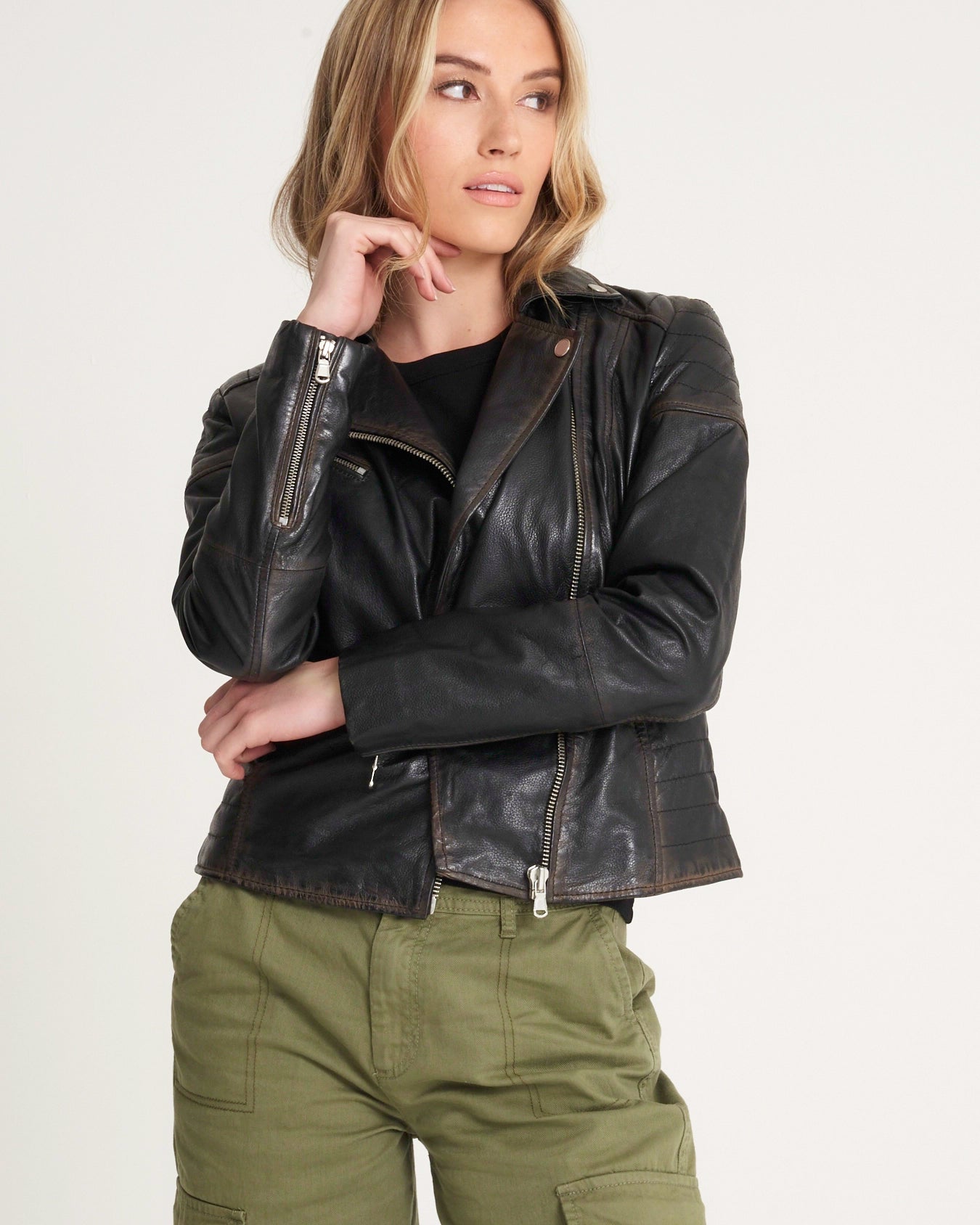 A woman wearing a black leather moto jacket with silver zippers, styled with olive green cargo pants against a plain light gray background.