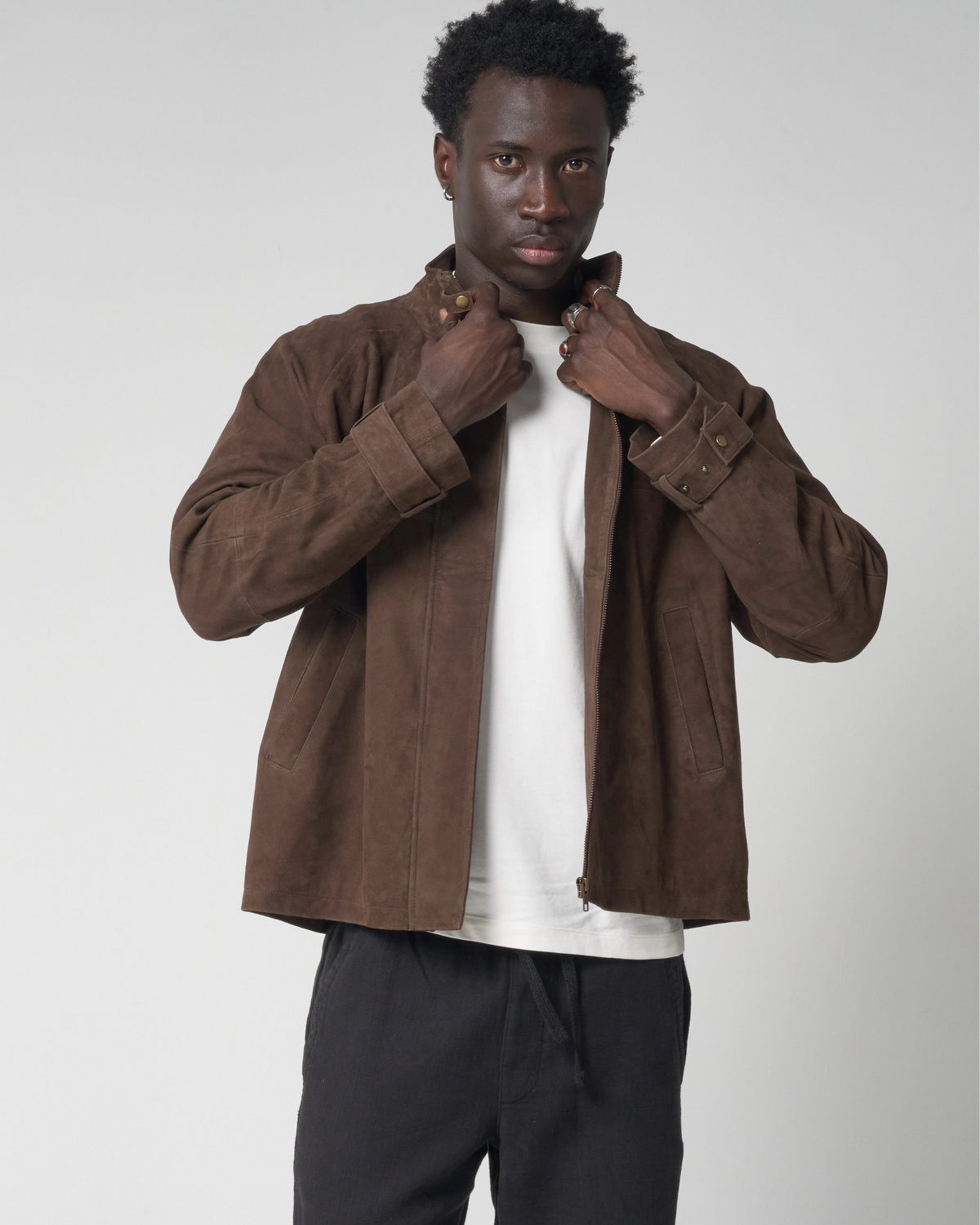 A man wearing a brown suede jacket over a white t-shirt and black sweatpants, standing against a light gray studio background.