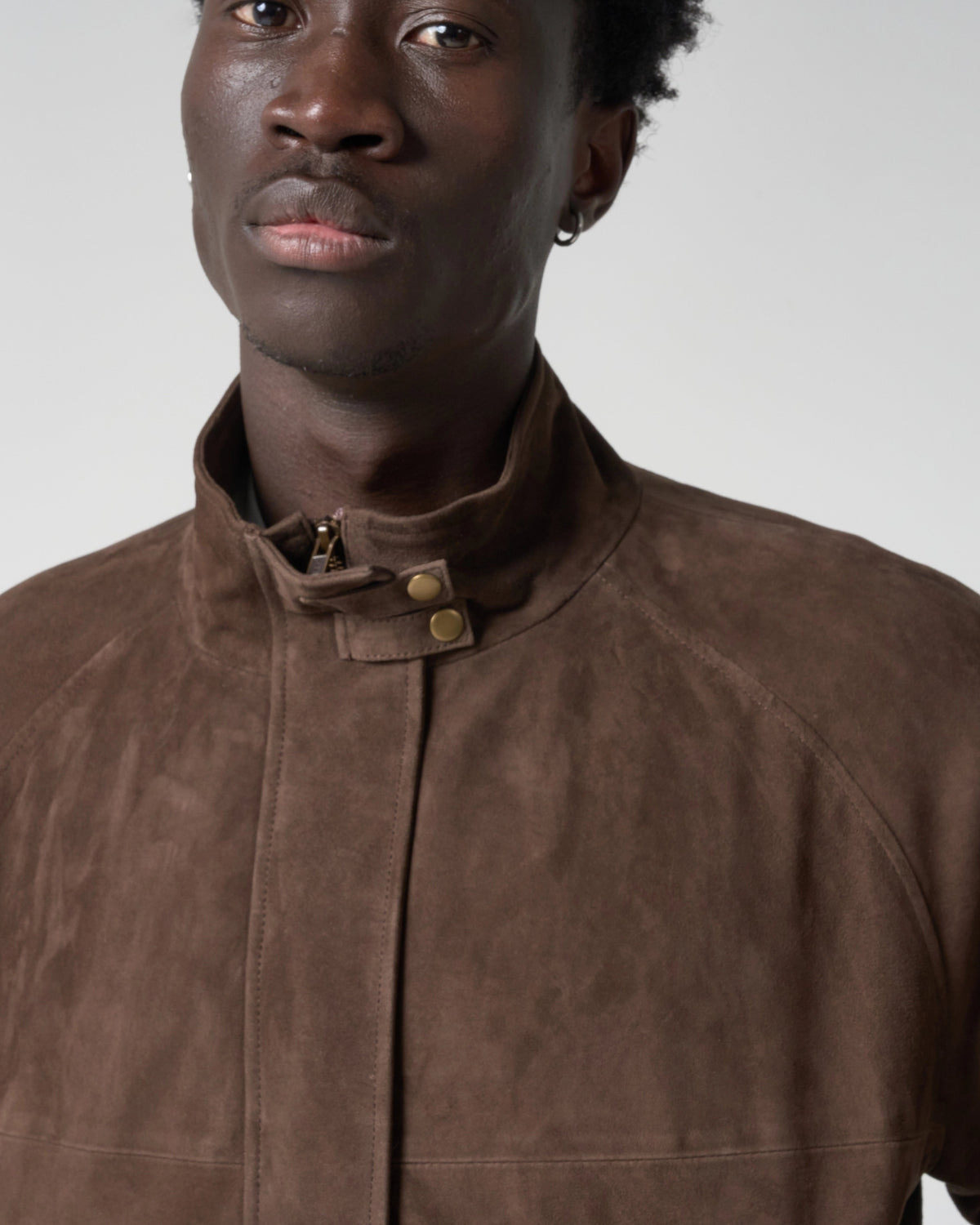 A close-up of a man wearing a brown suede jacket with a high stand-up collar and brass snap buttons, set against a plain light gray background.