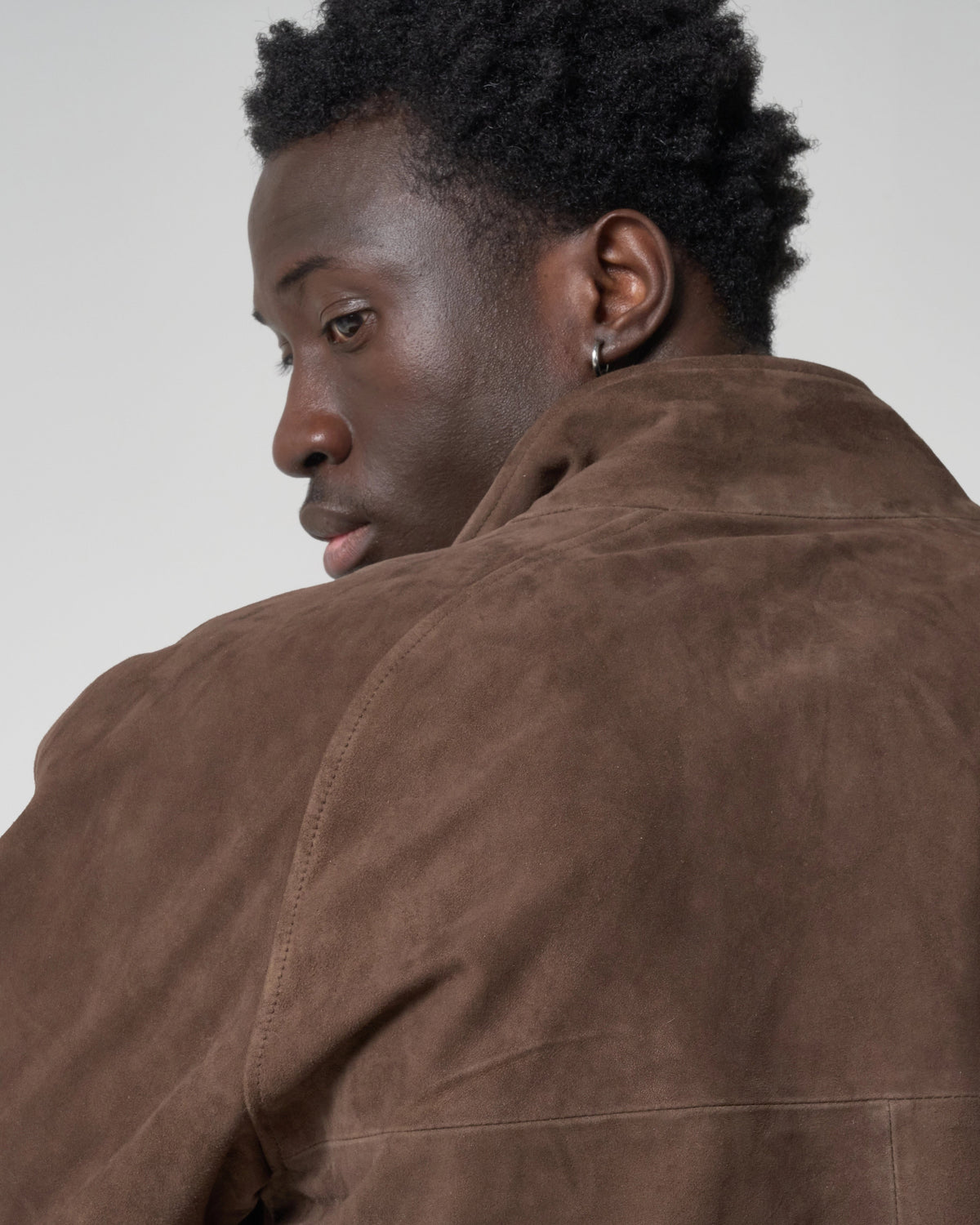 A close-up side profile of a man wearing a brown suede jacket against a light grey studio background.