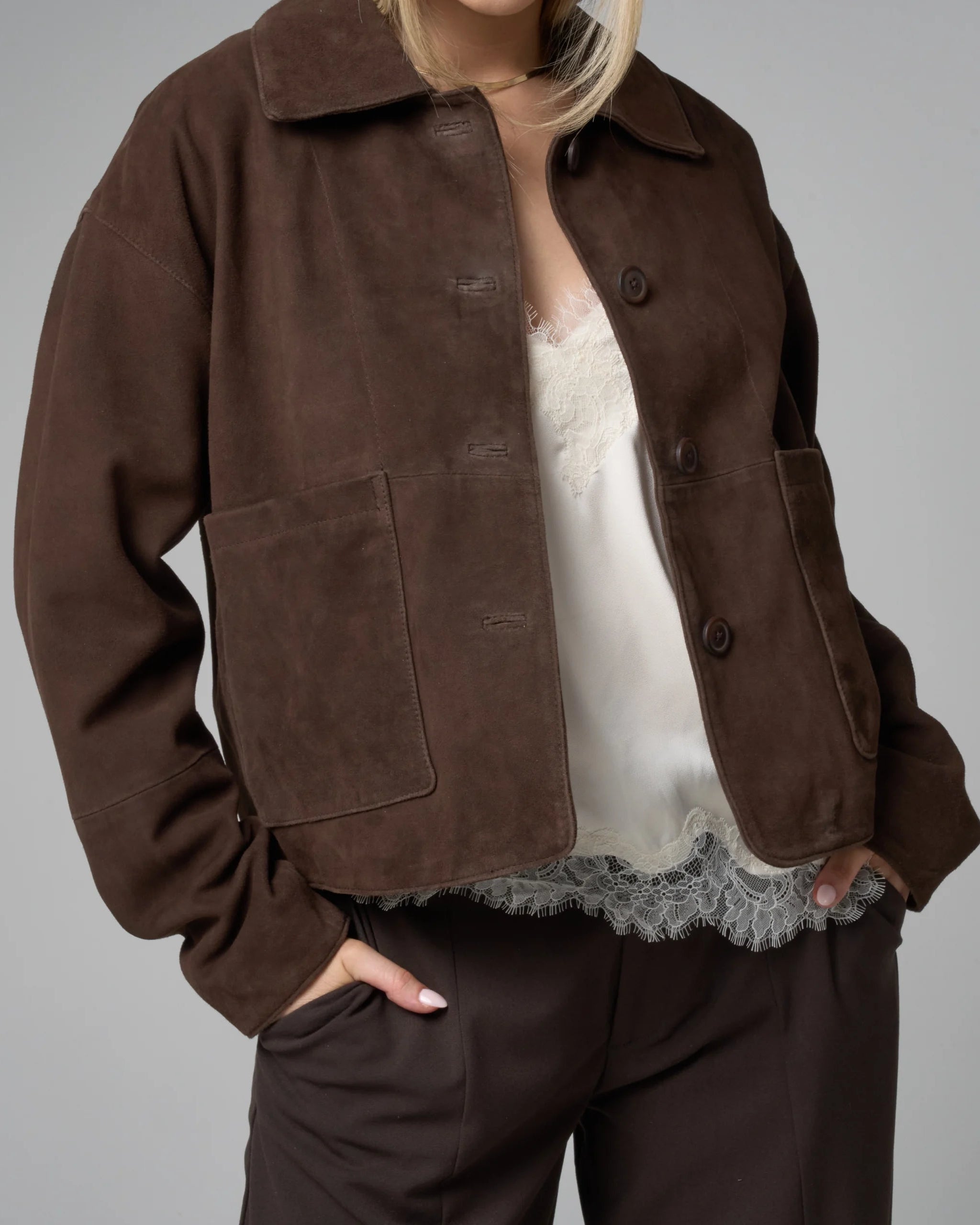 Person wearing a brown jacket over a white lace top on a gray background