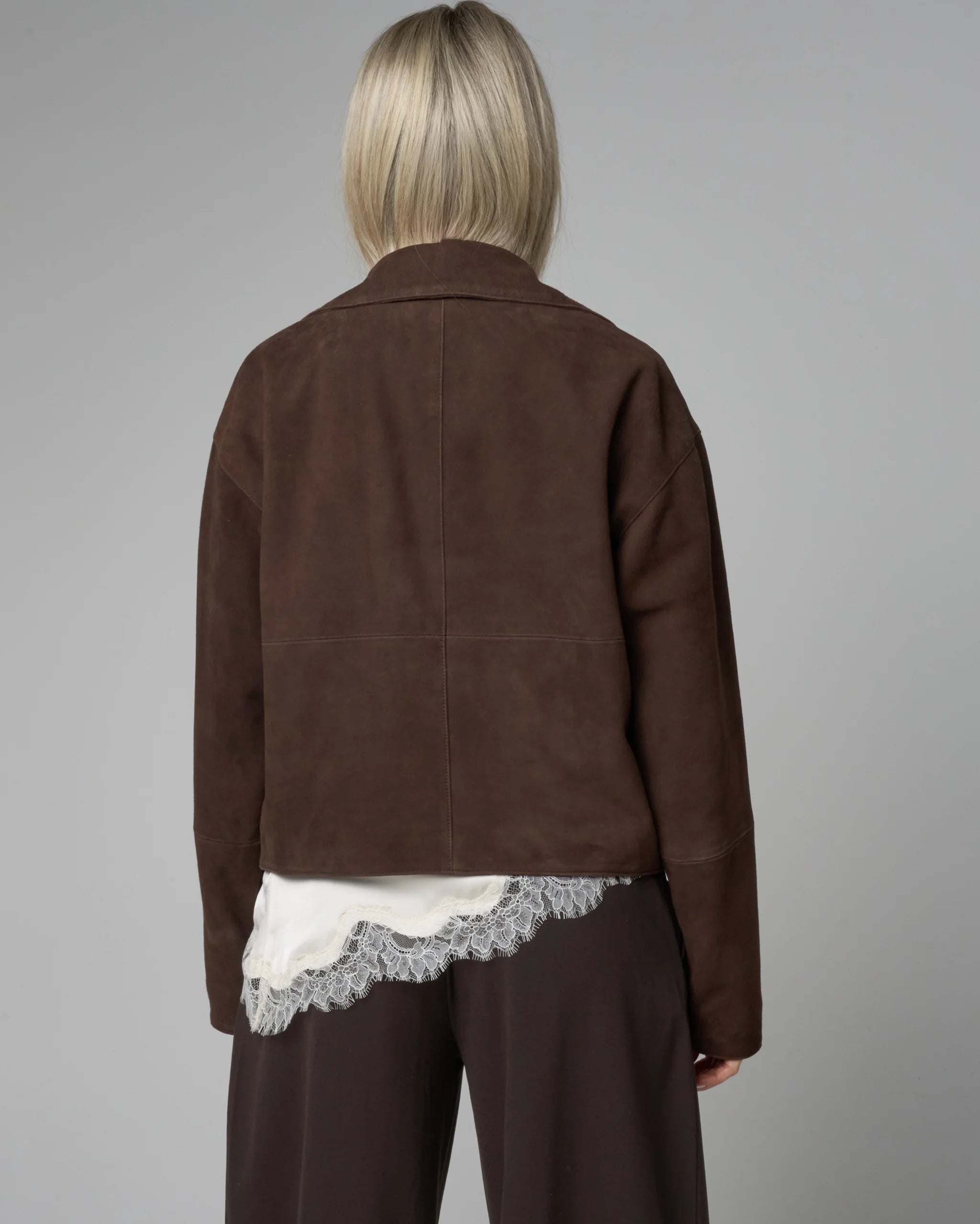 Person wearing a brown suede jacket over a white lace garment on a gray background. No logos or pattern. Cropped fit.