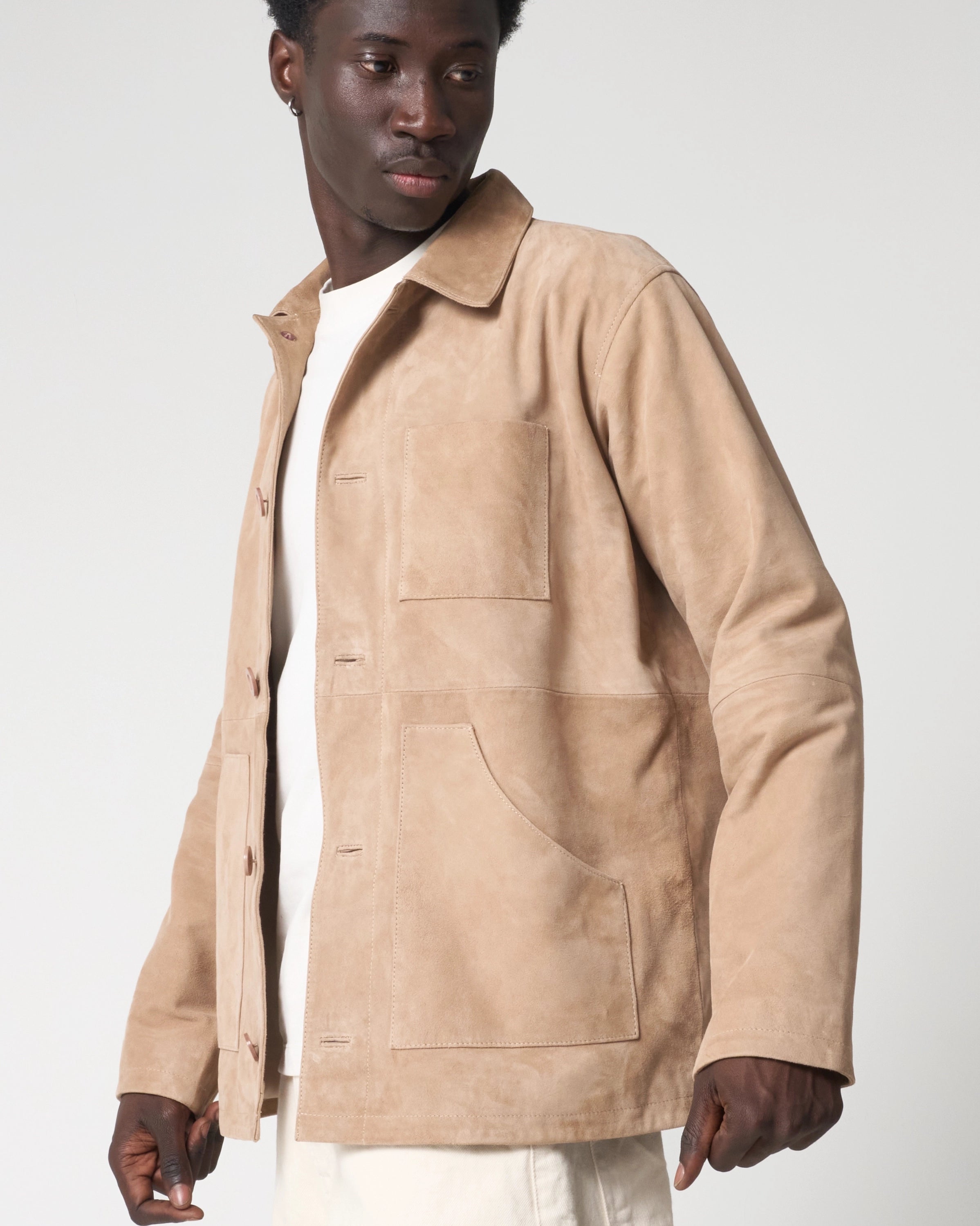 Side view of man styling the sandy beige suede chore jacket by Barneys Originals. The jacket has a collar, long sleeves, upper chest pocket and lower waist welt pocket. The jacket is worn unbuttoned.