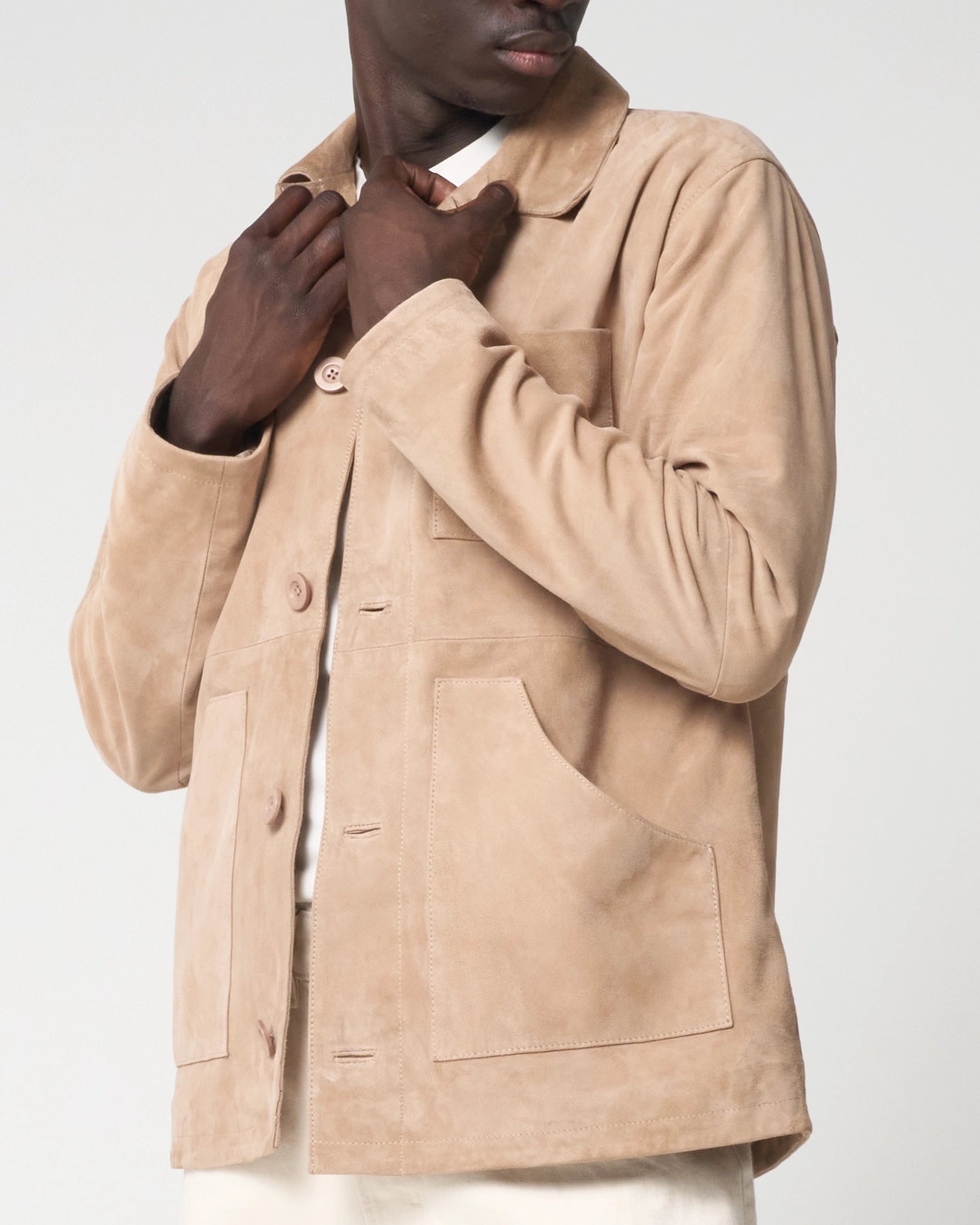 A close-up of a model wearing a beige suede jacket, adjusting the collar with both hands. The jacket features a buttoned front, a collar, and large front patch pockets.