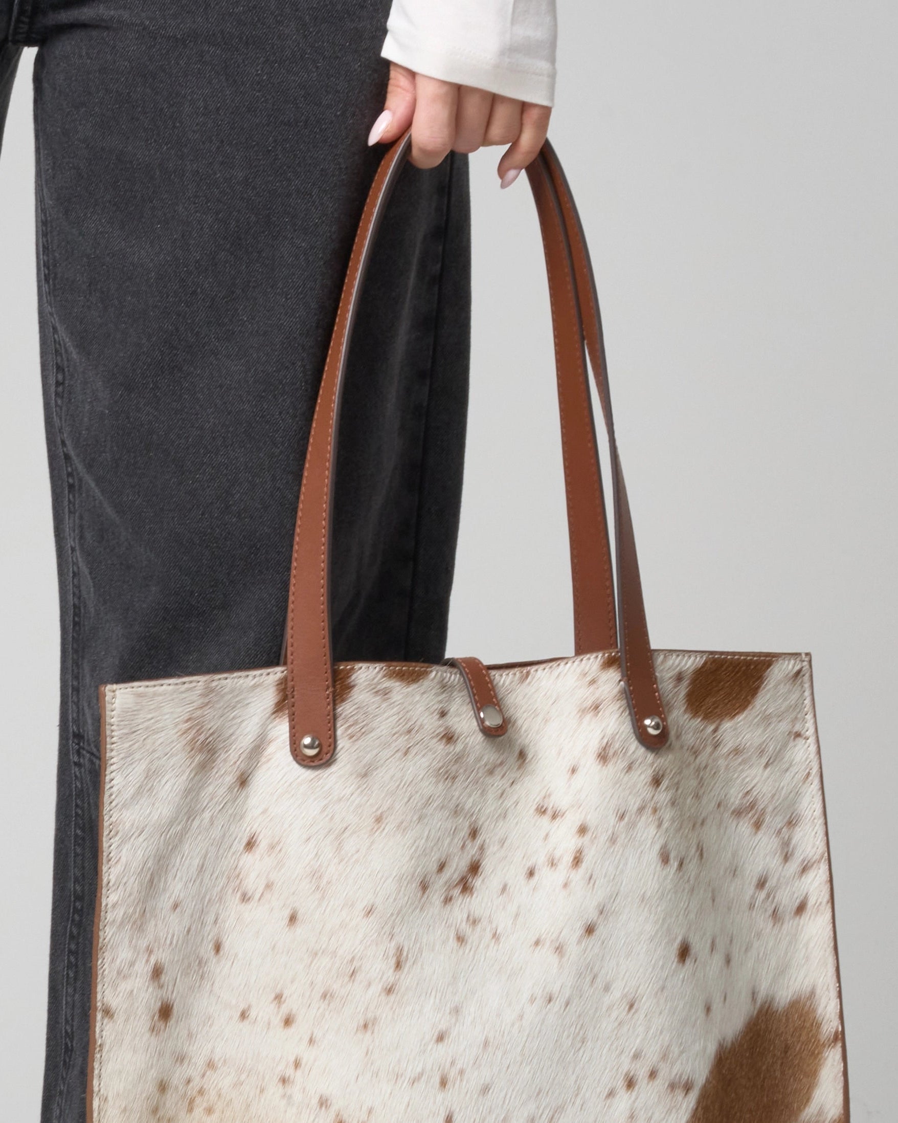 Person holding a tote bag with cowhide leather pattern and brown handles.
