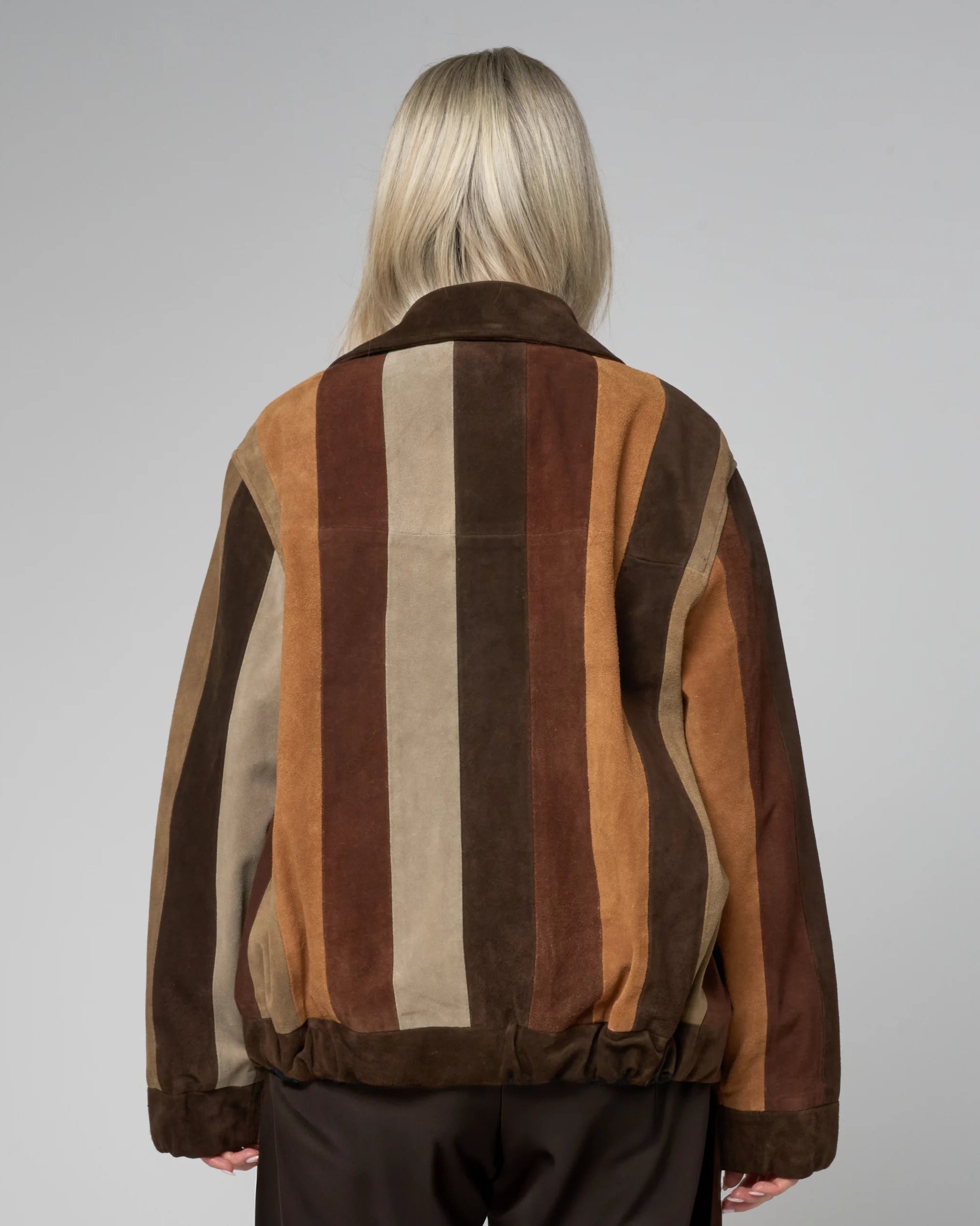 Back of a person wearing a multicolored striped suede jacket on a grey background. The stripes are beige, an and brown, running vertically.