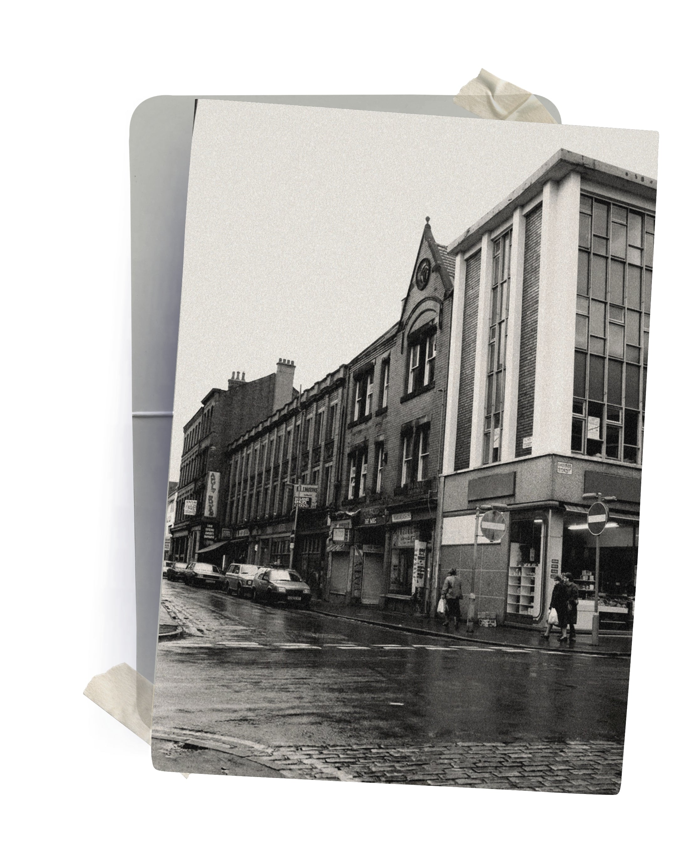 A square art print or postcard featuring a black and white photograph of a wet city street lined with brick and modern buildings, shown standing against a white background with a ribbon bookmark visible at the top.