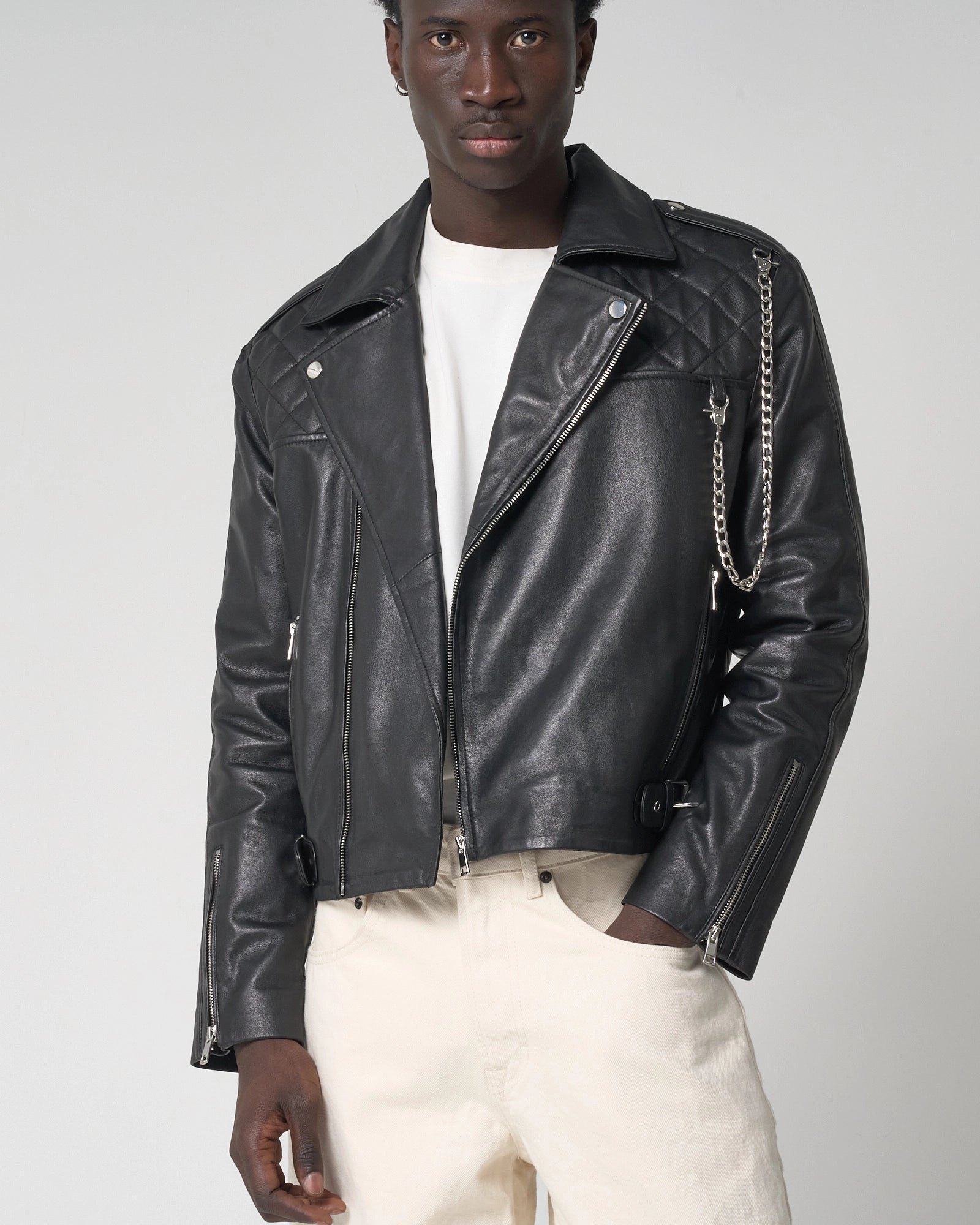 A man wearing a black leather moto jacket with silver chain accents, a white t-shirt, and cream-colored trousers, photographed against a neutral light gray background.