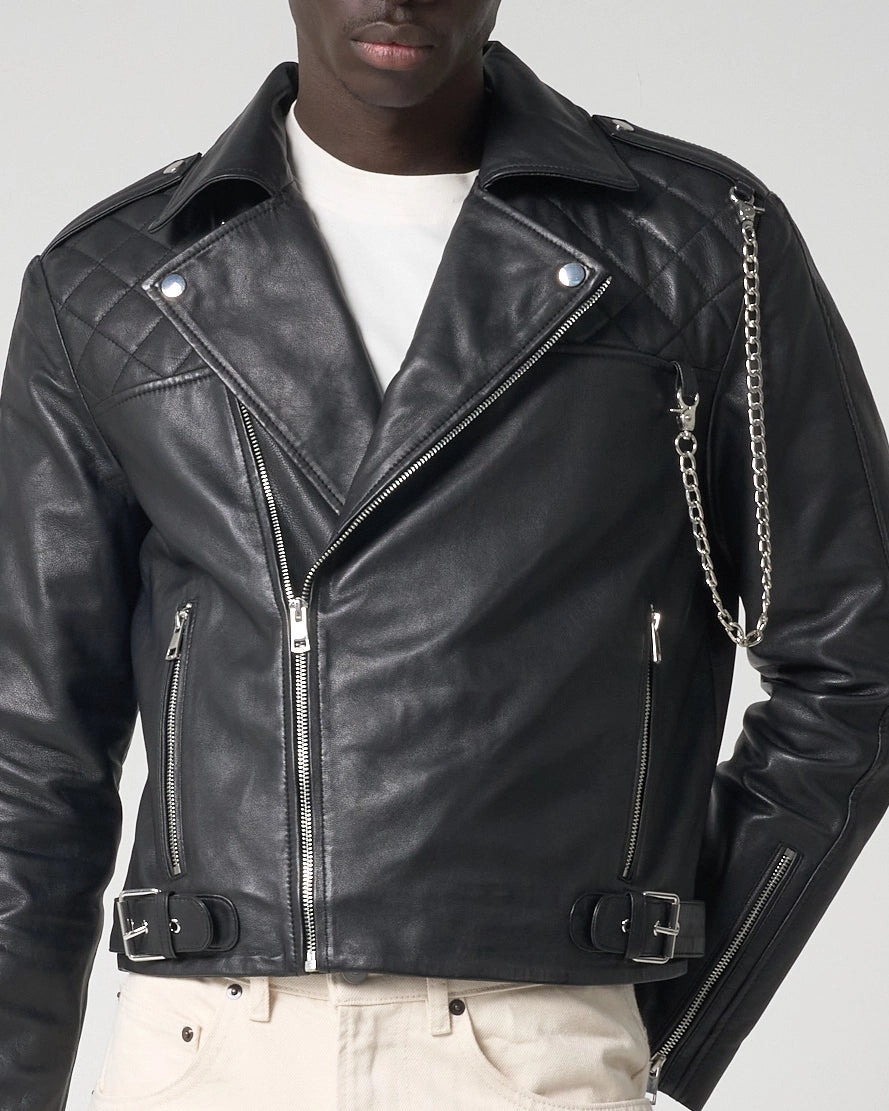 A close-up of a model wearing a black leather motorcycle jacket with quilted shoulder panels, silver hardware, and a silver chain detail on the shoulder, styled over a white t-shirt and white pants.