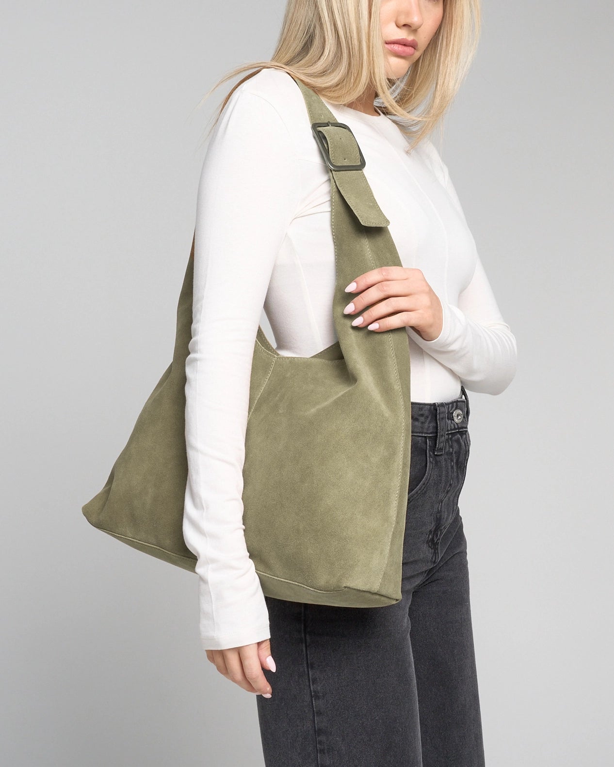 A woman wearing a white long-sleeve top and dark grey jeans carries an olive green suede slouchy shoulder bag with a wide adjustable strap against a light grey photo studio background.