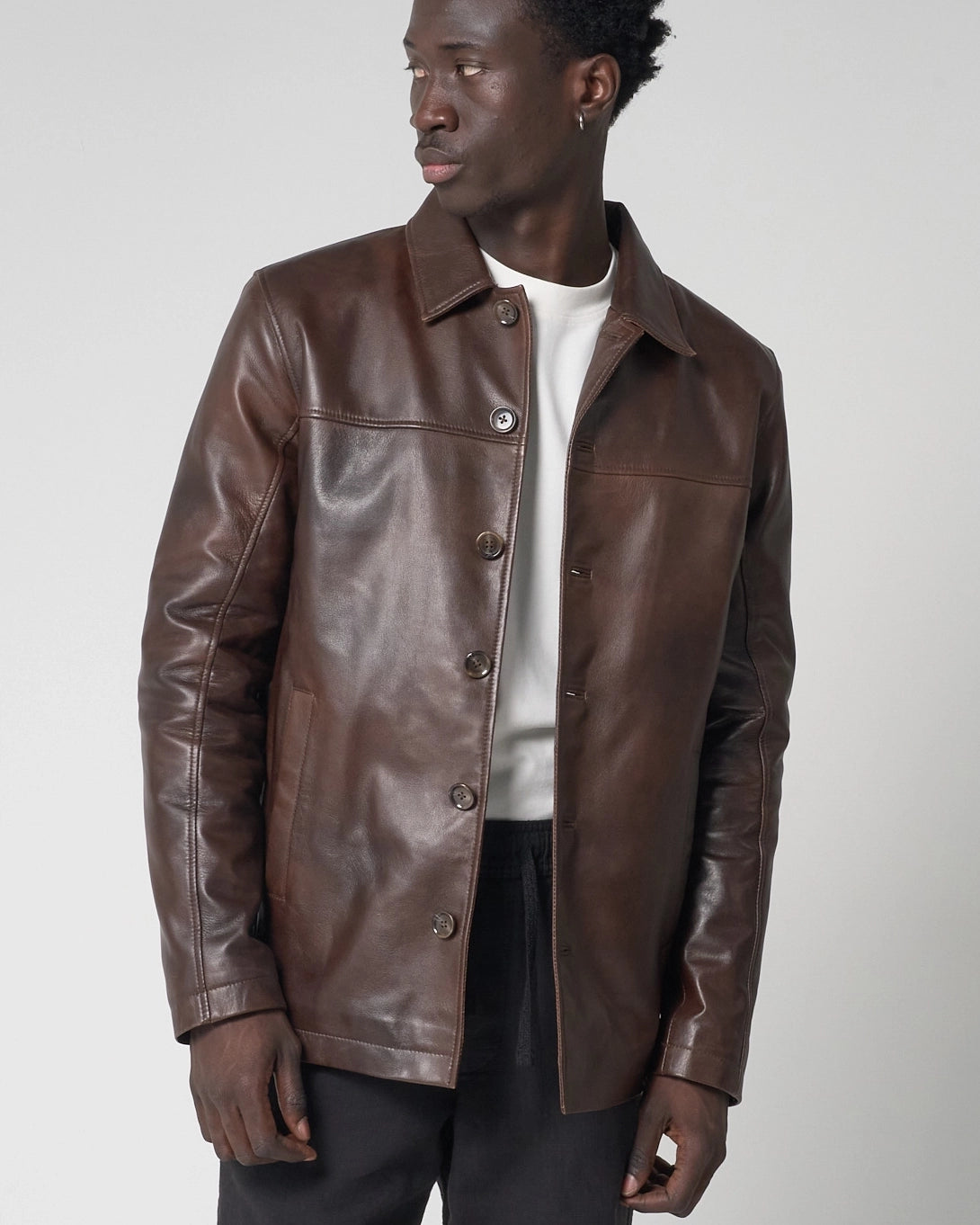 A man wearing a brown leather jacket with buttons and a collar. The jacket is worn unbuttoned to expose the white tshirt below.